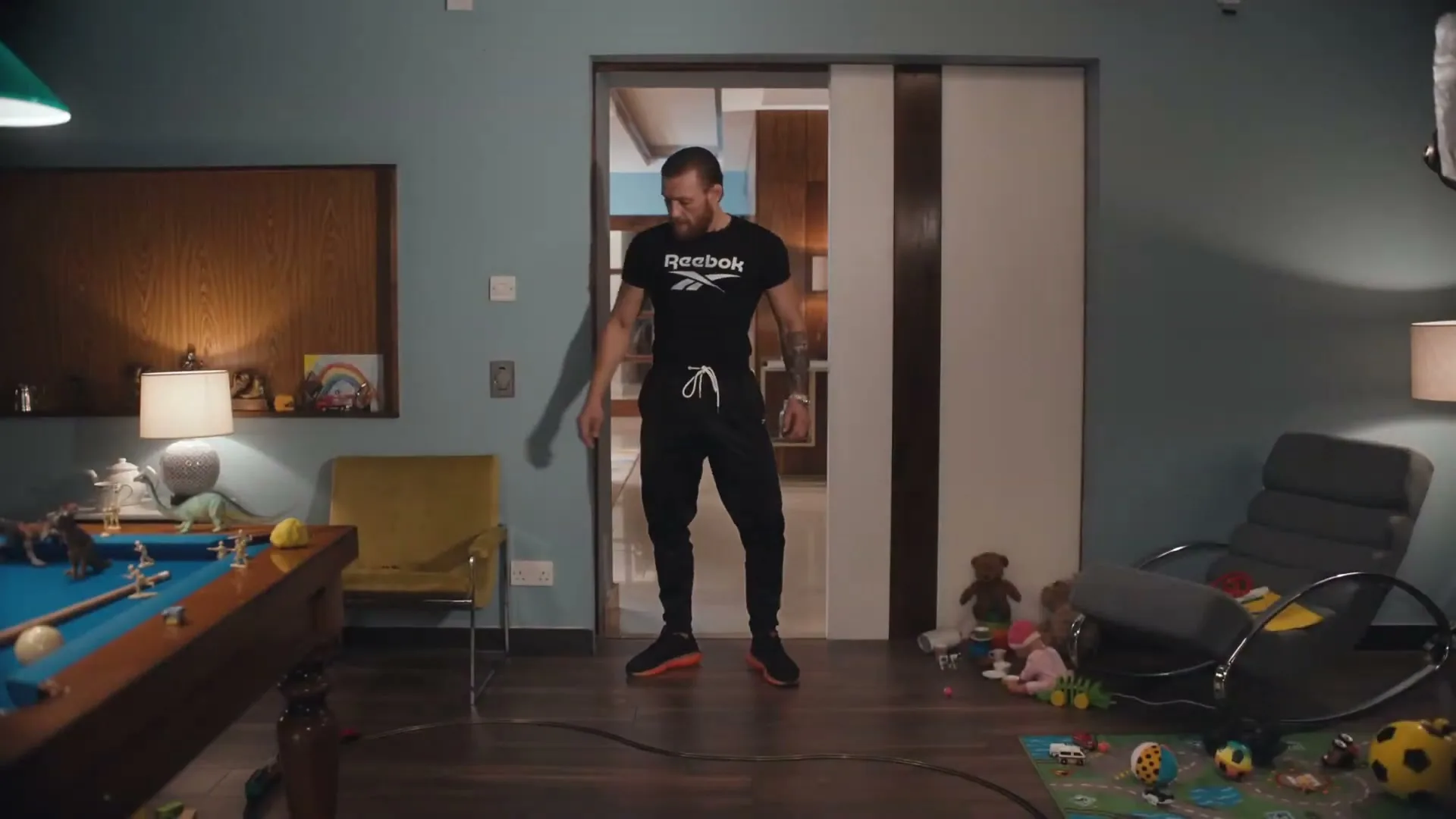 A man in a black Reebok t-shirt and sweatpants stands in a messy living room with a pool table and toys, looking around.
