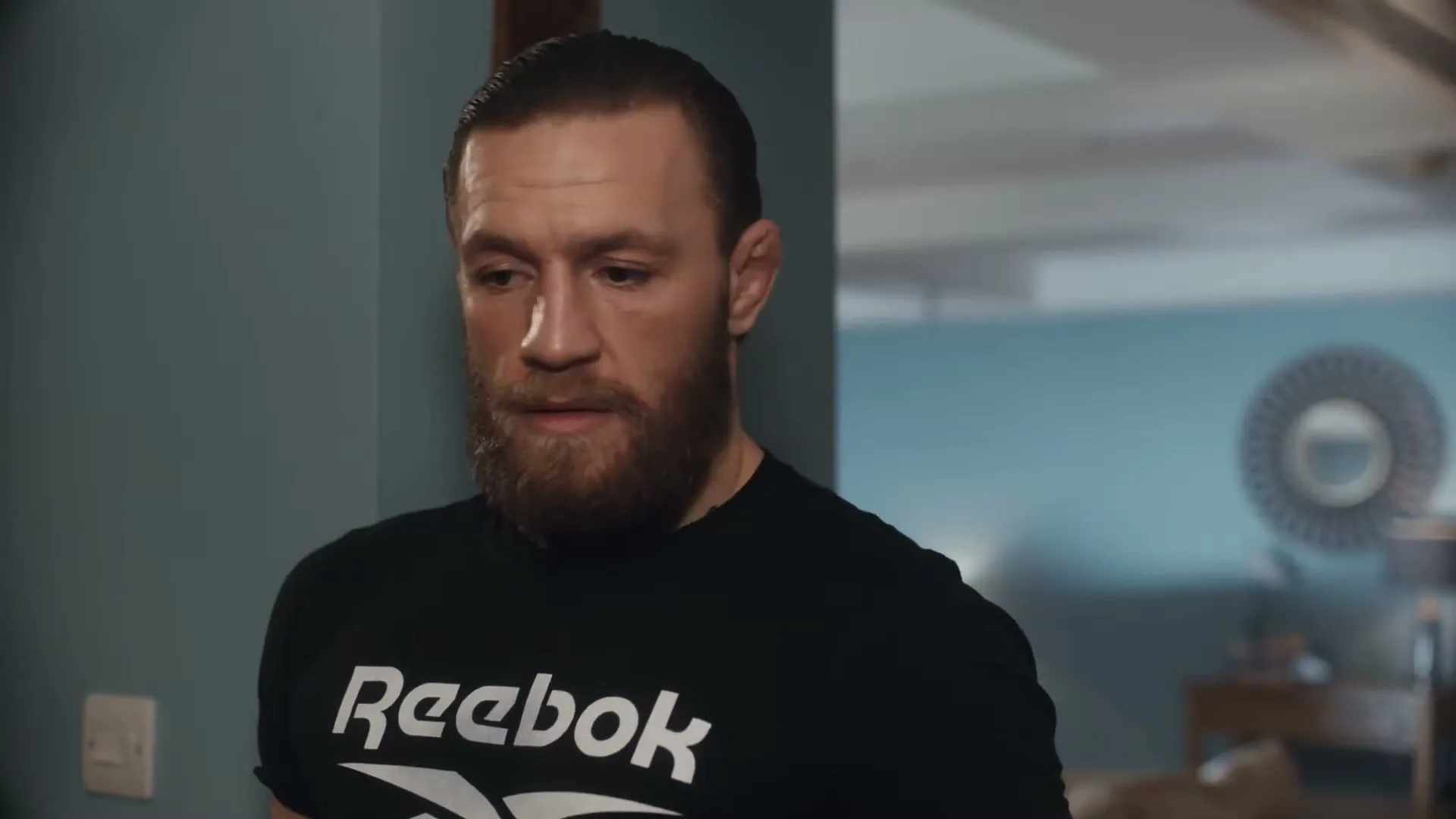 Conor McGregor, a bearded man in a black Reebok t-shirt, looks down and then to the side in a modern living room.