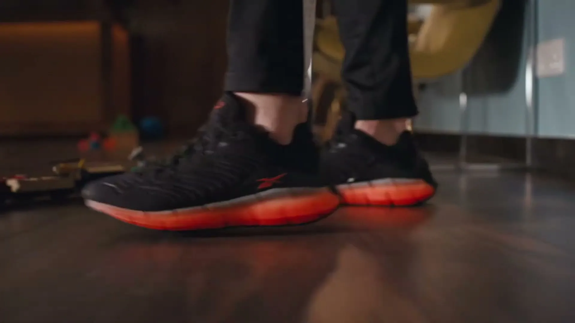 A person wearing black and orange Reebok sneakers steps on a toy train, causing sparks to fly, on a wooden floor in an interior setting.