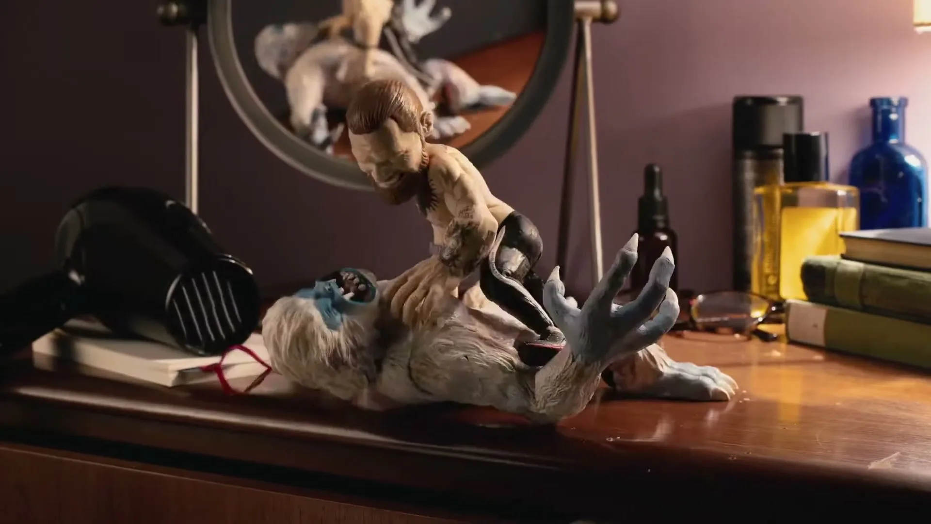 A stop-motion animation shows a small, bearded male action figure in black shorts wrestling a large, furry yeti-like creature on a wooden dresser in a bedroom.