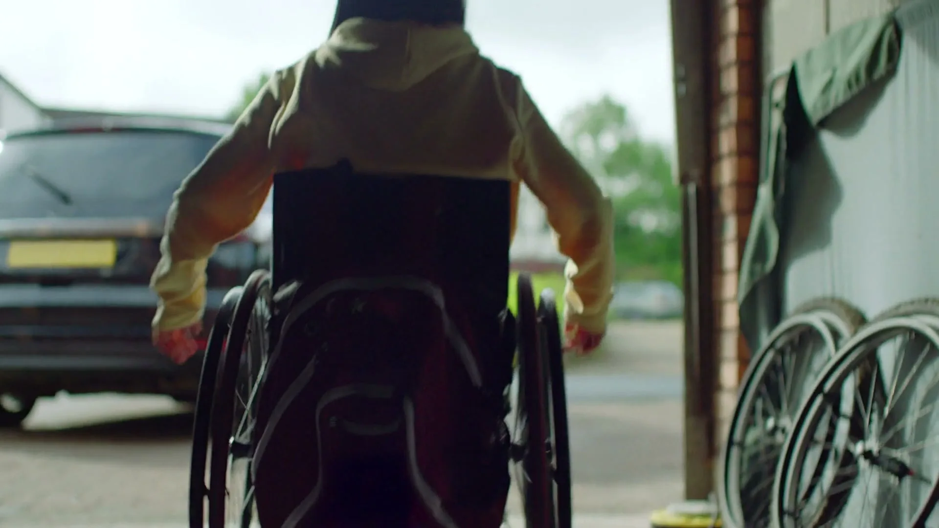 A person in a wheelchair with a backpack on their back, wearing a light yellow hoodie, is seen from behind, moving out of a garage into a driveway.
