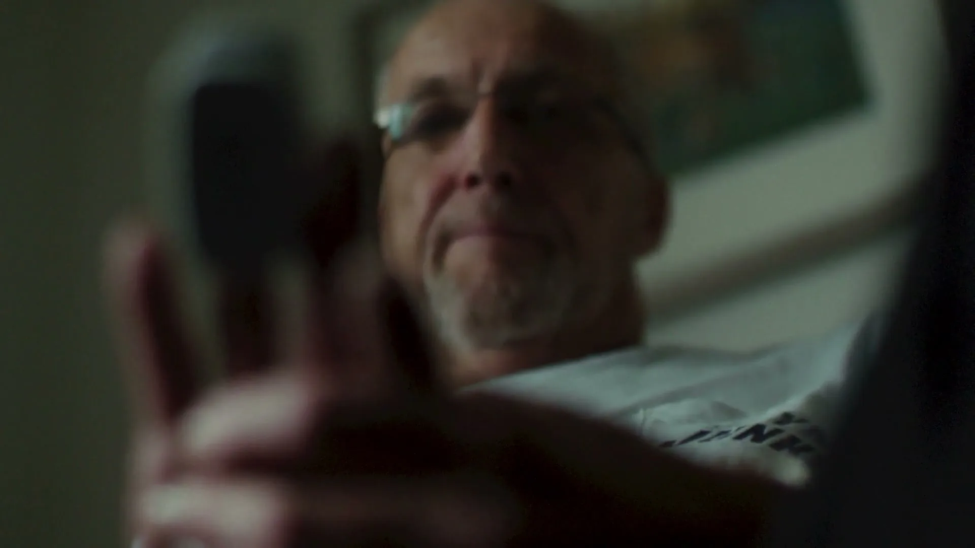 A man with glasses and a beard holds a pulse oximeter on his finger, looking at the device in an interior setting.