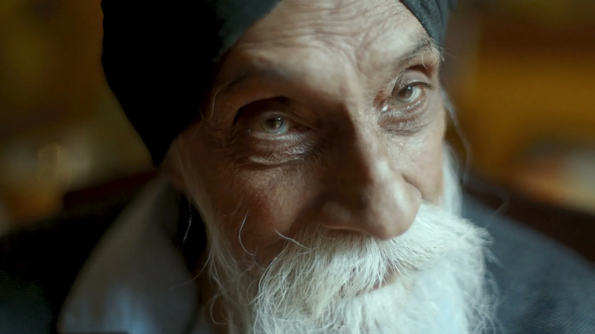 A close-up of an elderly man with a white beard and black turban, looking directly at the camera with a slight smile.