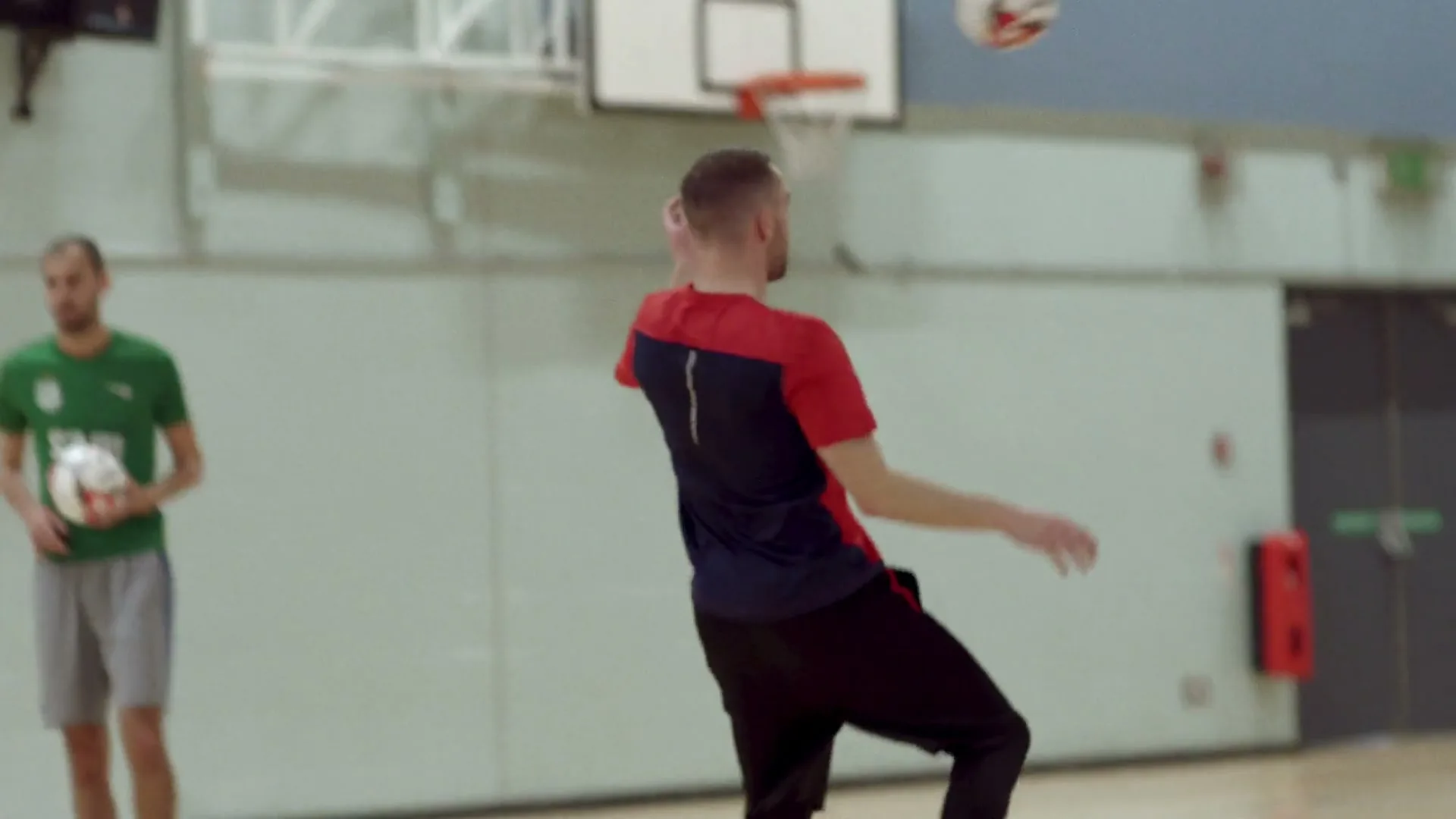 A man in a red and blue shirt plays basketball in a gym, while another man in a white shirt stands in the foreground.