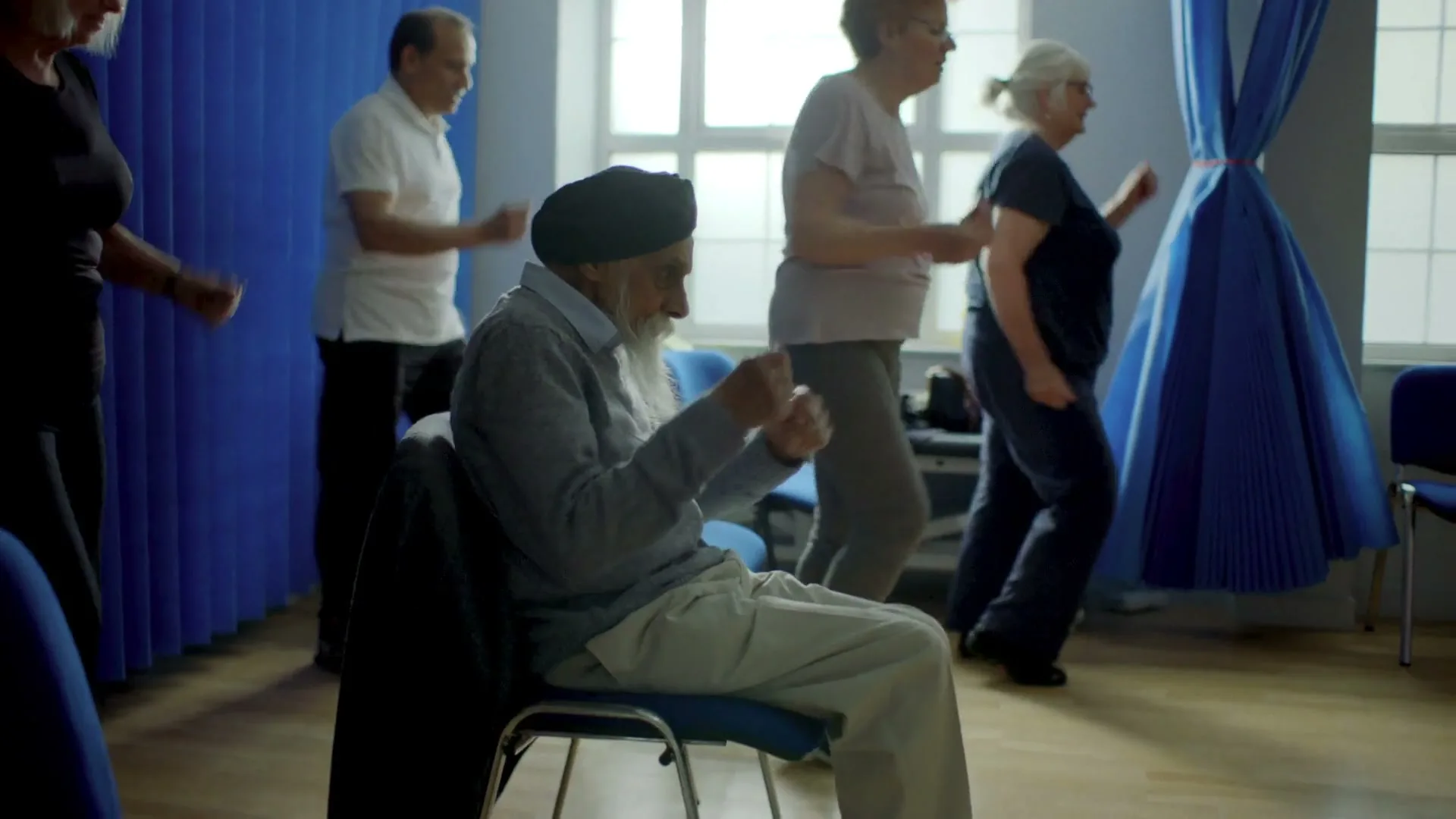 A diverse group of older adults, including a man in a turban, participate in a seated exercise class in a bright room with blue curtains.
