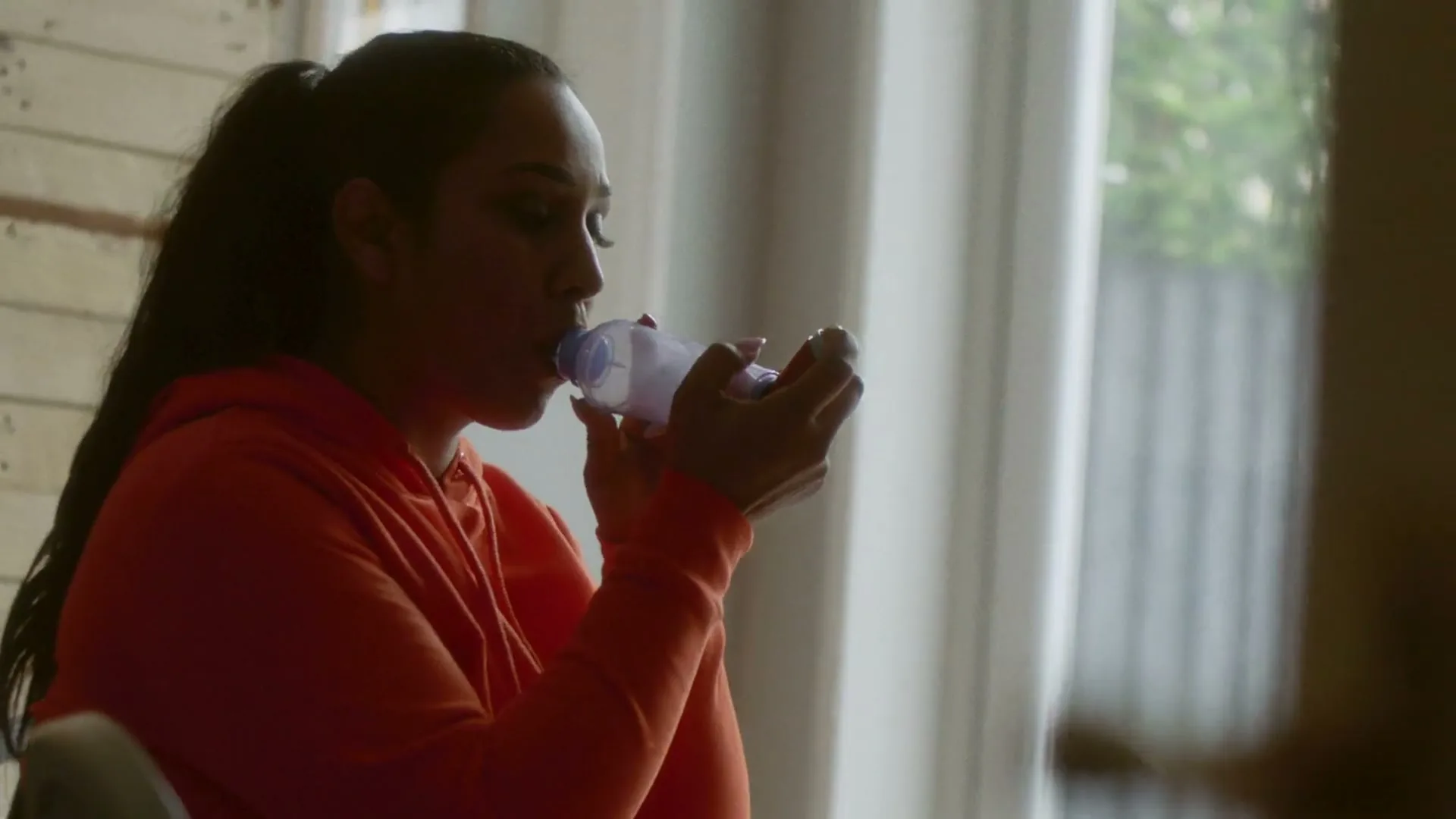 A woman with dark hair in a ponytail, wearing an orange hoodie, uses an inhaler with a spacer in a sunlit room.
