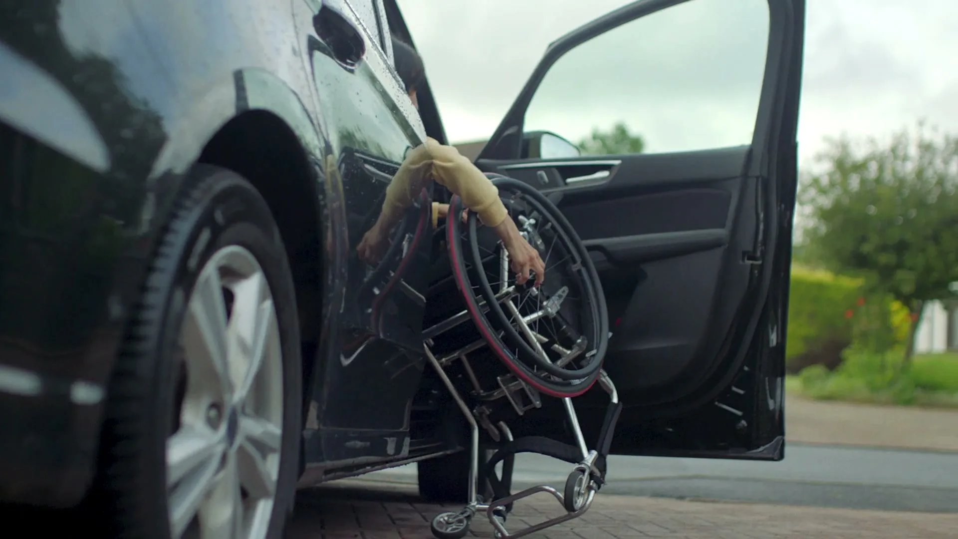 A person with a disability struggles to get their wheelchair out of the back of a black car on a cloudy day.