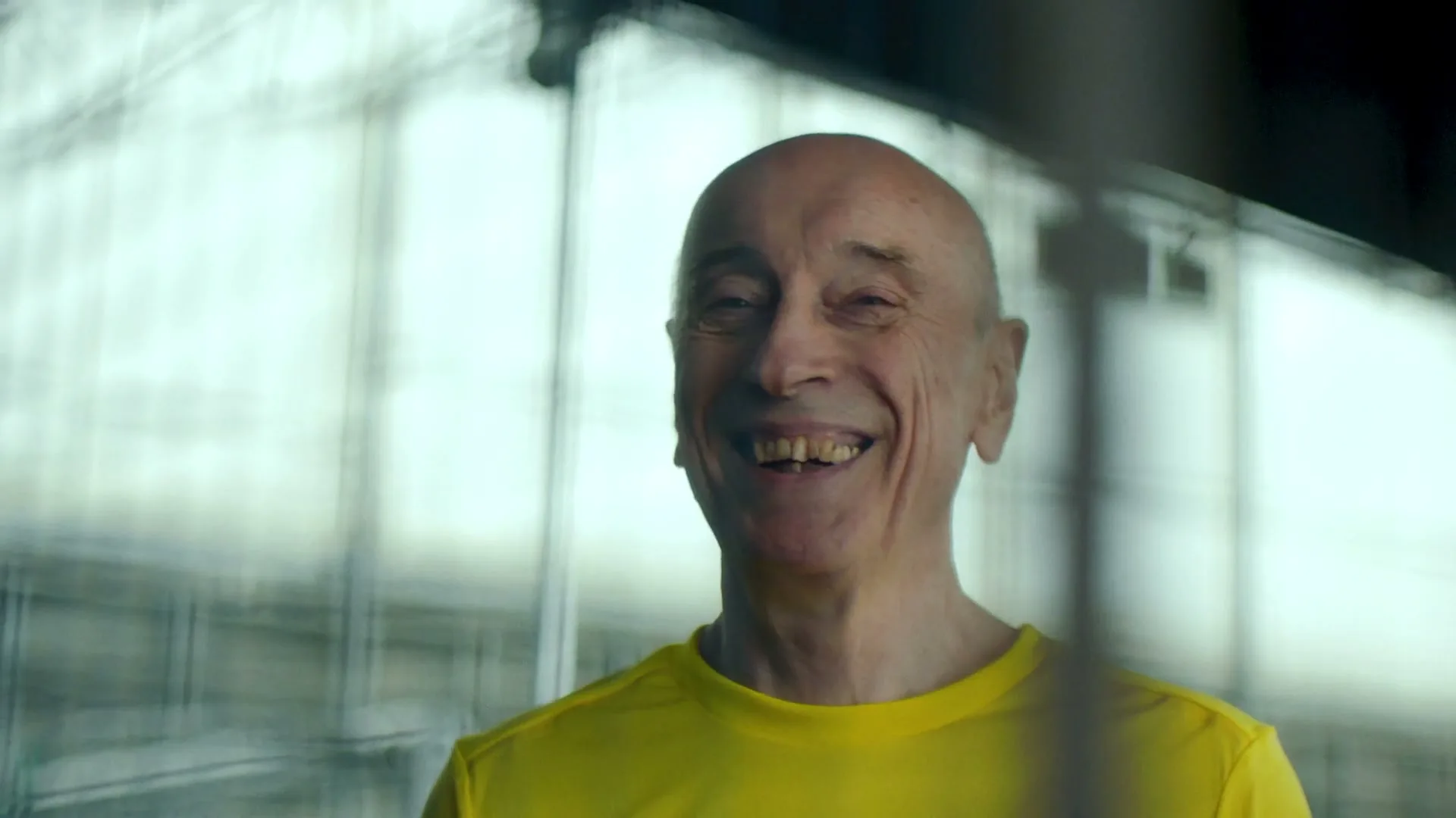 A bald man in his 60s, wearing a yellow t-shirt, smiles with his eyes closed, in an interior setting with a blurred background.