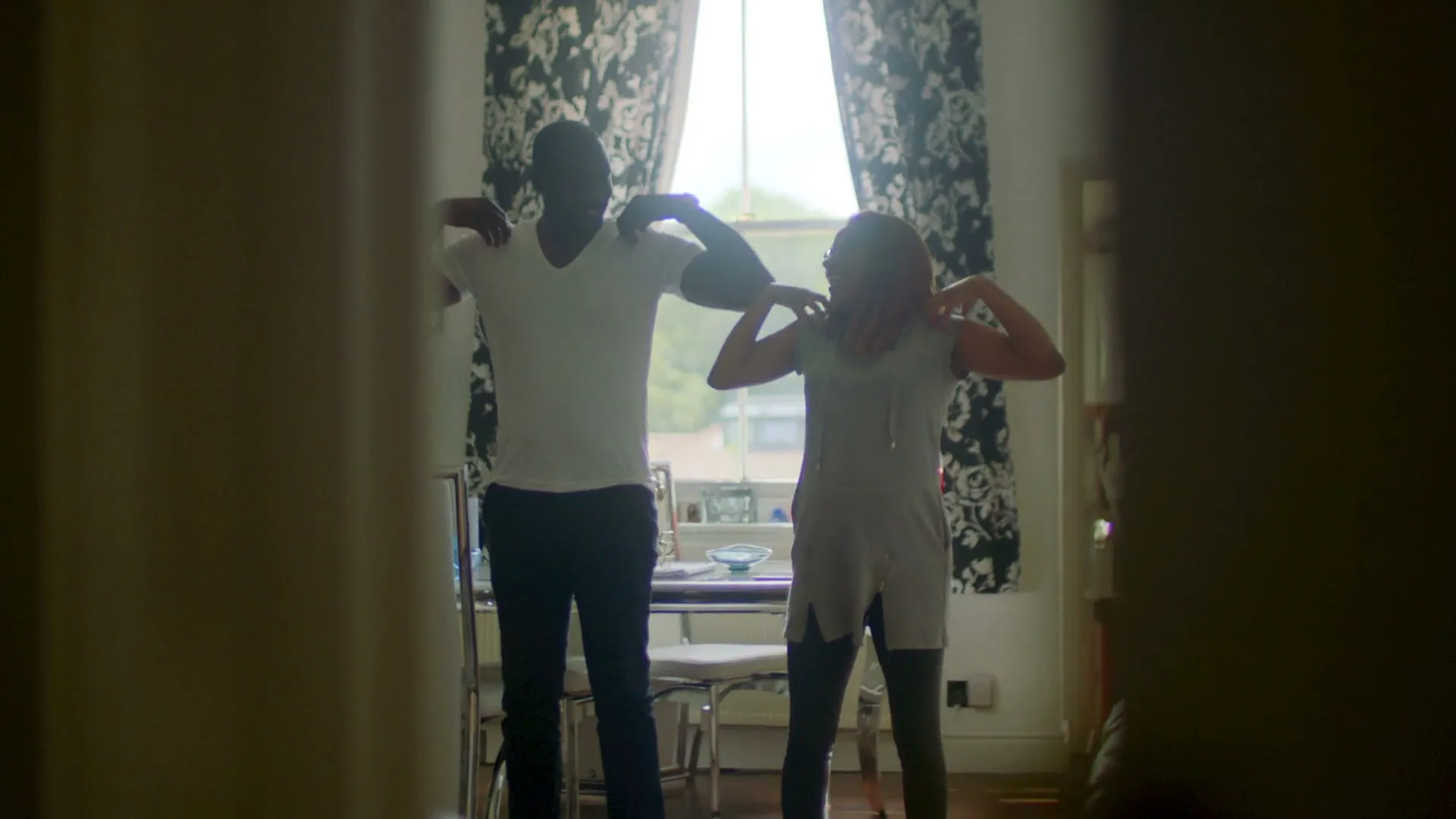 A man and a woman, both silhouetted, stand in a bright room, stretching their shoulders, seen through a doorway.