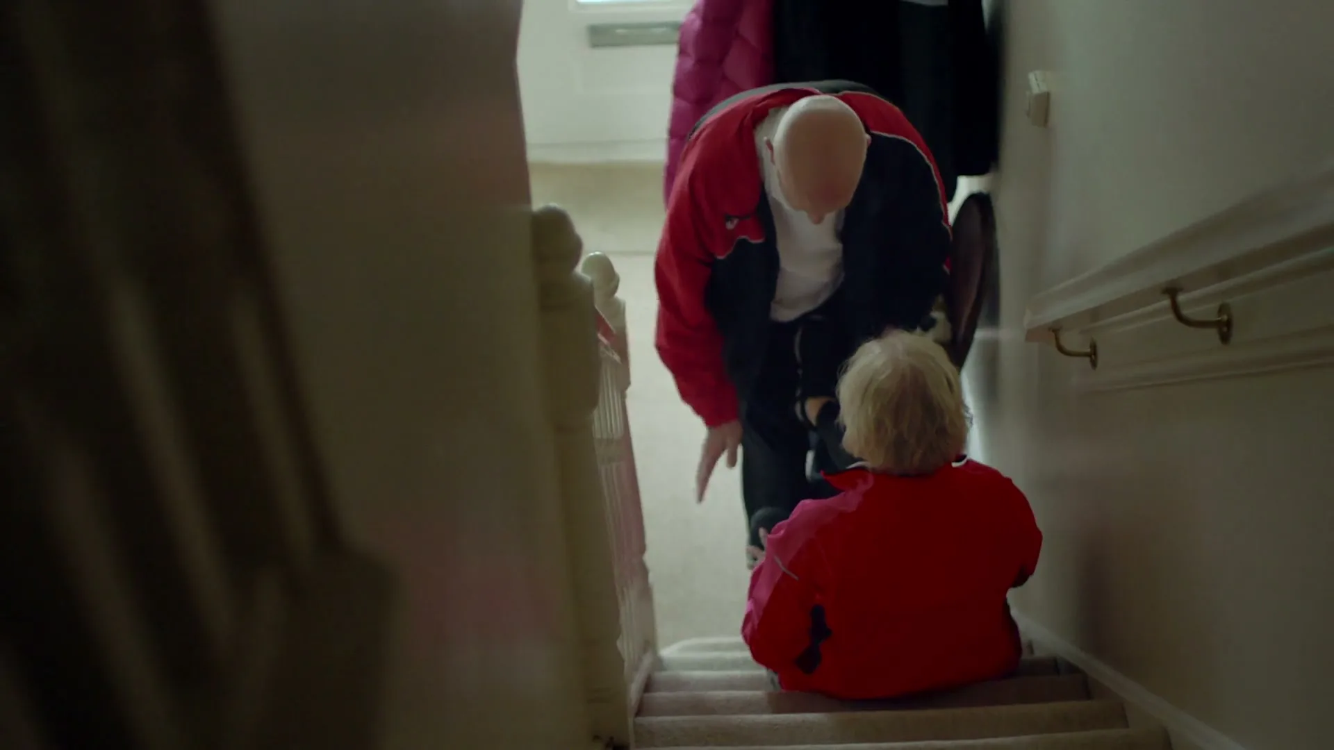 An older man in a red jacket helps an older woman in a red jacket up the stairs in a house.