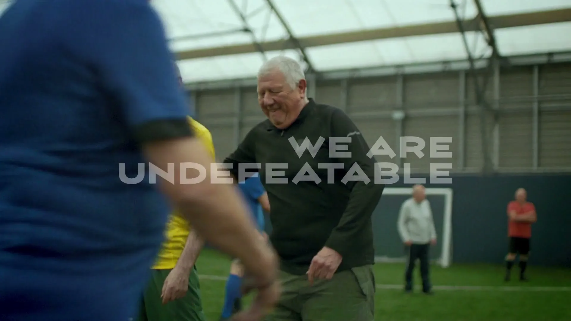 Two older men, one in a yellow shirt and one in a black shirt, walk and smile on an indoor soccer field with other players in the background.