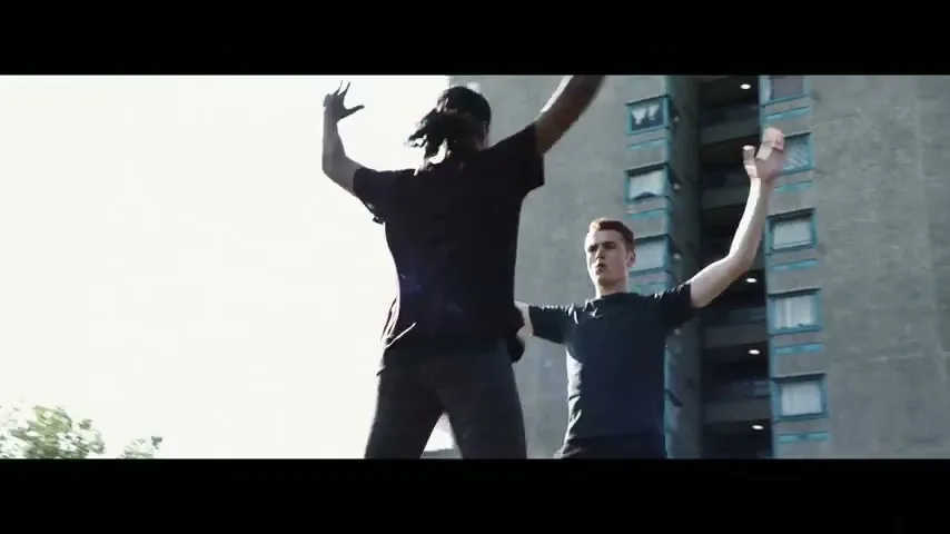 Two young men in athletic wear perform jumping jacks in front of a tall, grey apartment building under a bright sky.