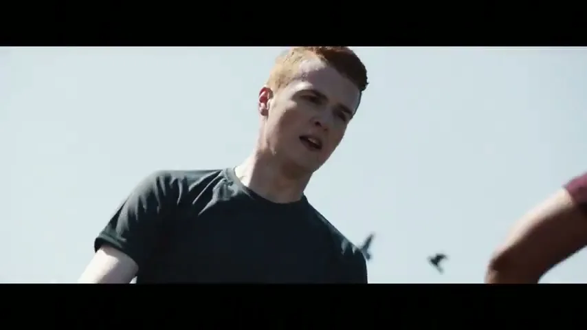 A young man with red hair in a black t-shirt looks intensely at another person, partially visible, against a bright sky.