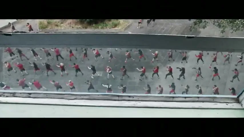 A man in a grey t-shirt looks out a window at a large group of people exercising outdoors, then the shot cuts to a closer view of people boxing.