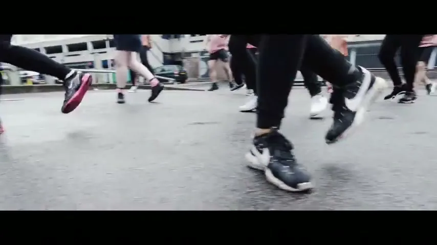 A low-angle shot captures the legs and feet of a diverse group of people running on a wet urban street.