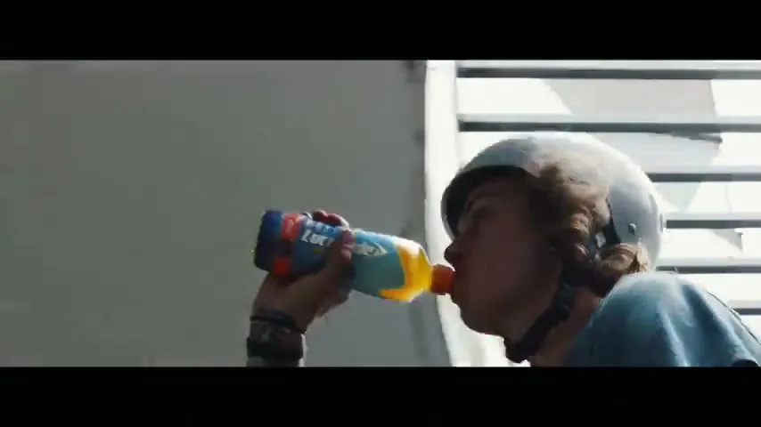 A young male skateboarder wearing a helmet drinks from a Lucozade bottle in an urban skatepark.