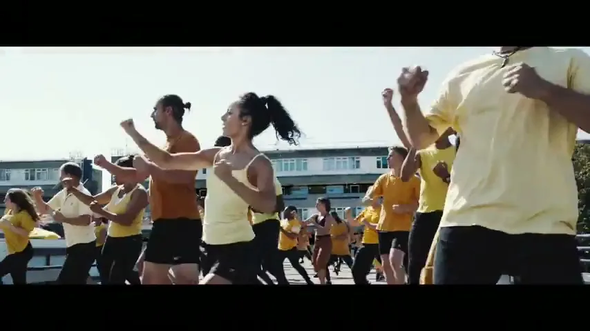 A diverse group of people in yellow and black activewear perform synchronized martial arts moves outdoors on a sunny day.