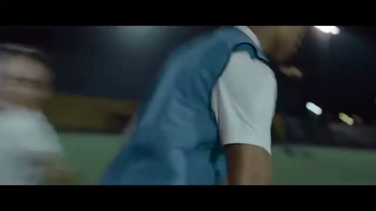 A male soccer player in a blue vest and white shirt kicks a soccer ball into a goal on a dark field at night.