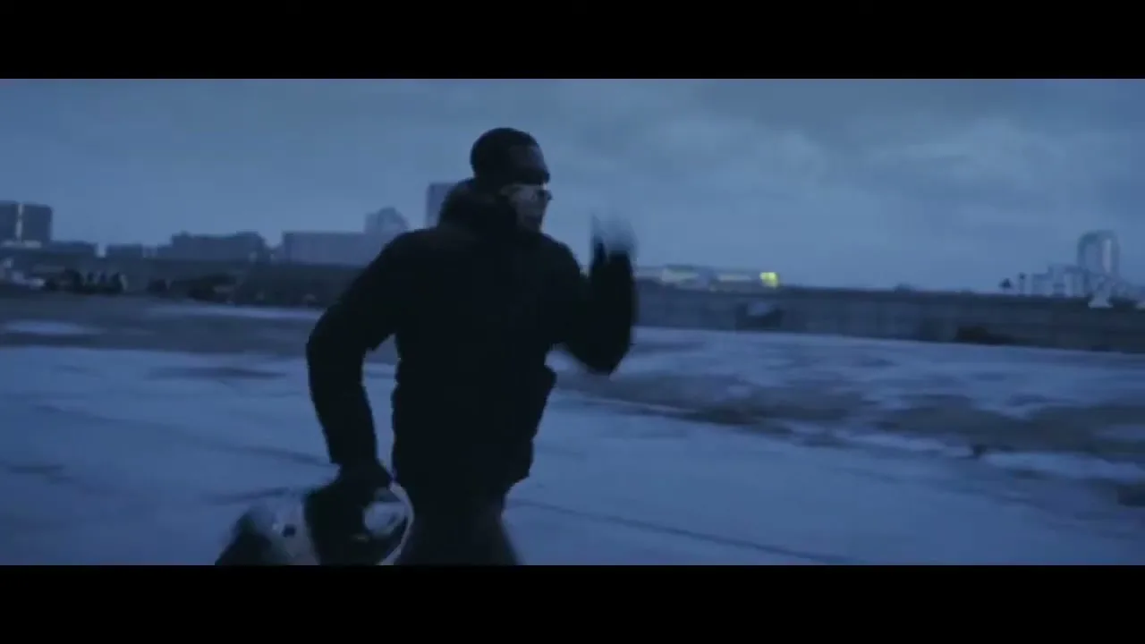 A male boxer in a dark hoodie and sunglasses runs across a desolate, wet, concrete area with a city skyline in the background under a dark, cloudy sky.