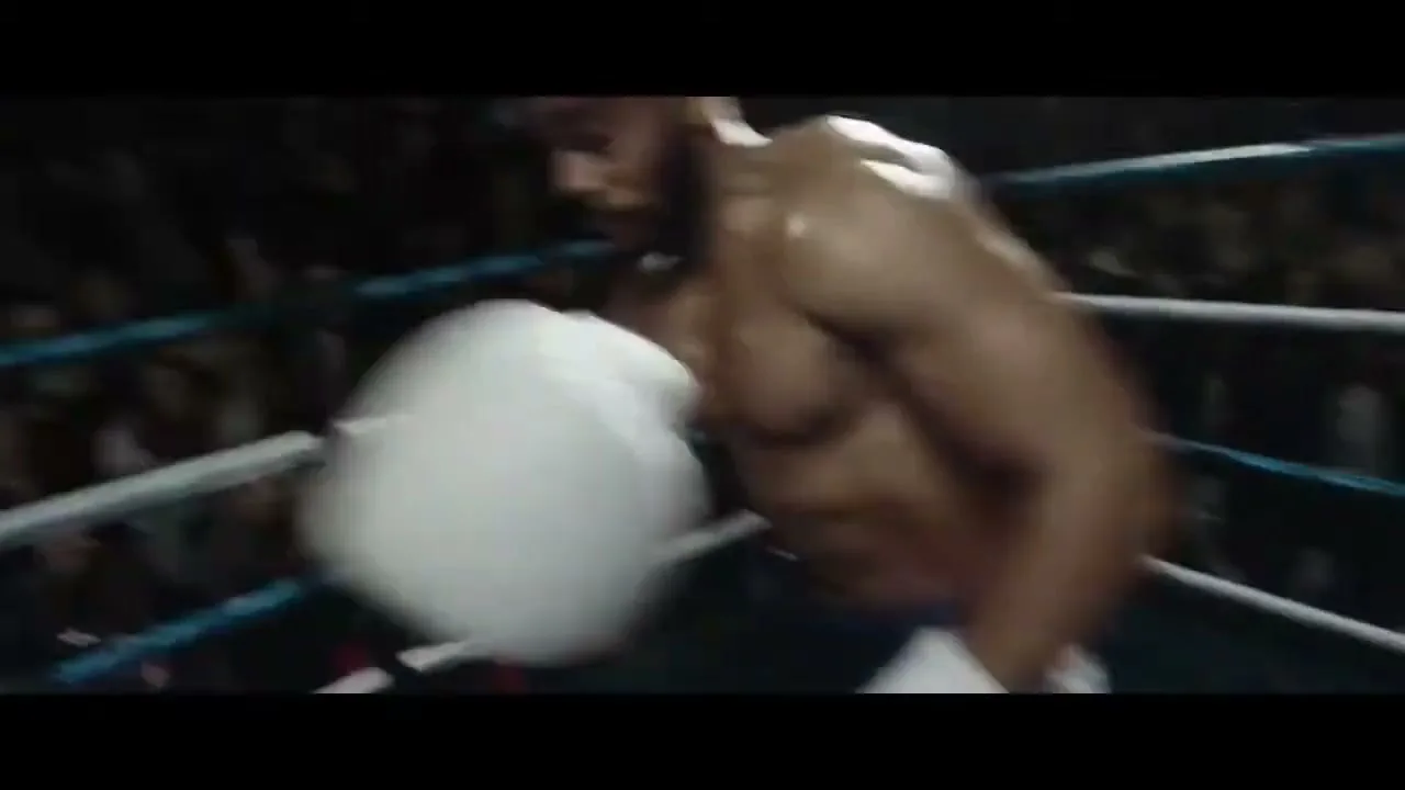 A male boxer with white gloves and black shorts throws a punch in a boxing ring, with a blurred crowd in the background.