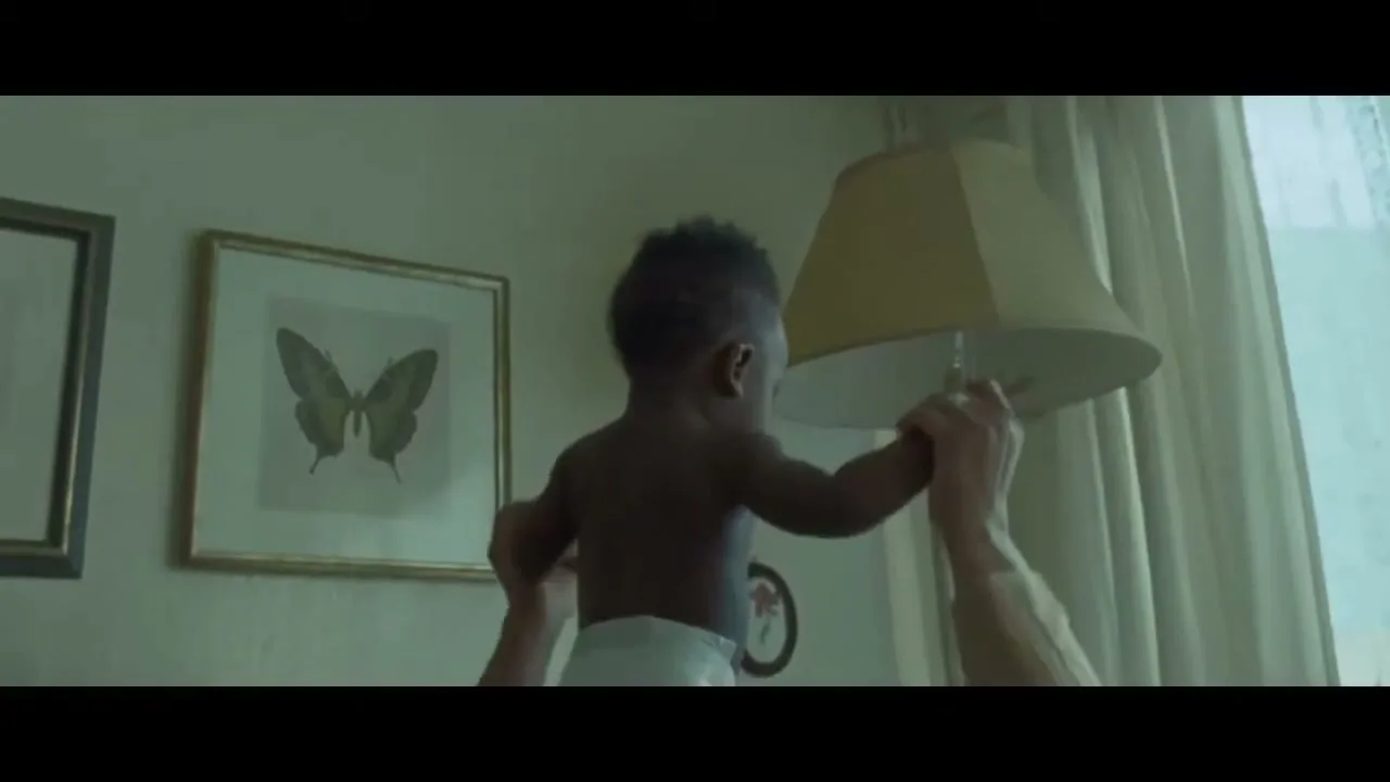 A baby in a diaper is held up by adult hands, reaching for a lamp in a room with framed butterfly art.