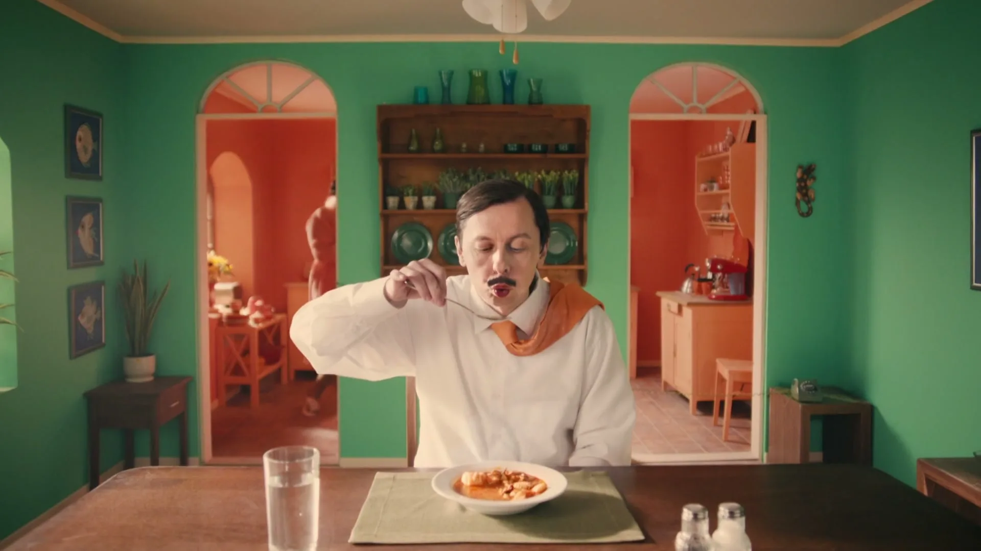 A man with a mustache and an orange scarf sits at a dining table, eating from a bowl with a spoon, in a vibrant green and orange room.