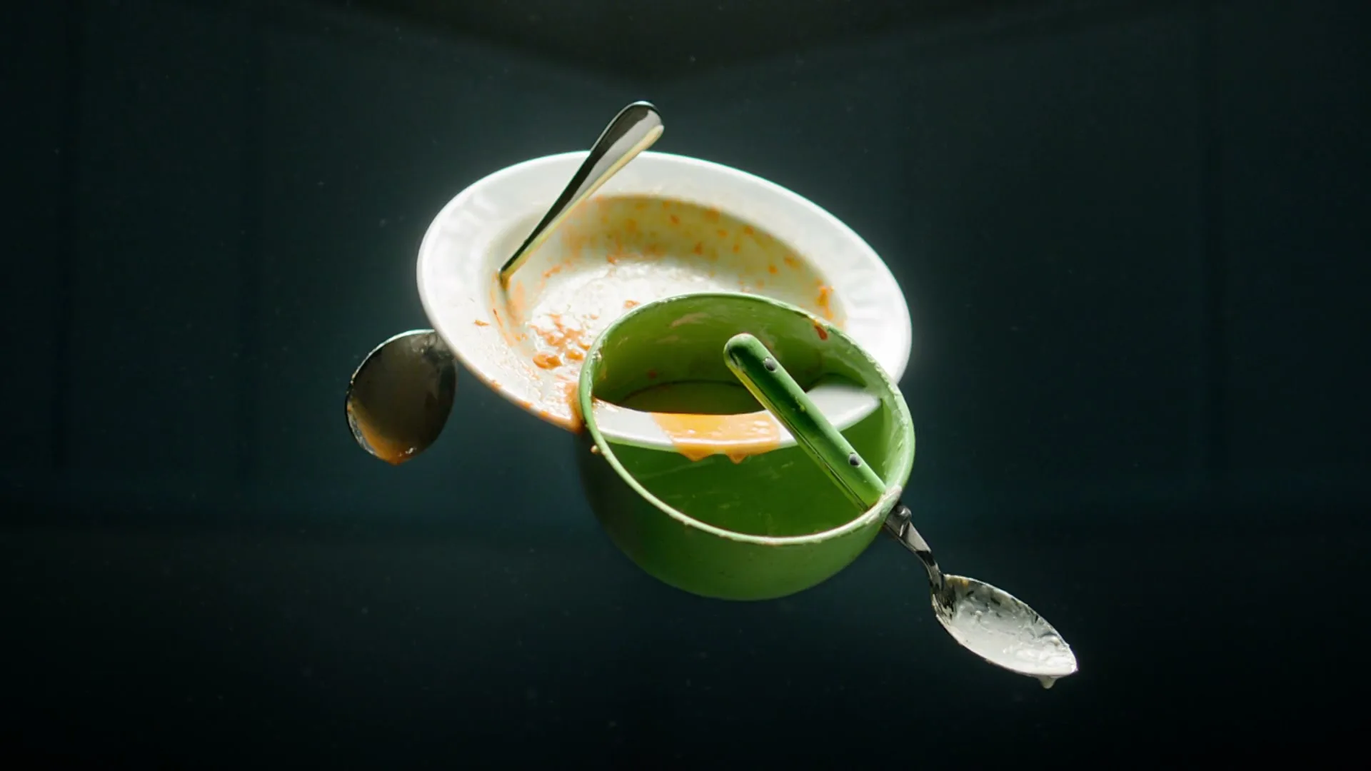 A white plate and a green bowl, both dirty with food residue, float in a dark void with spoons.