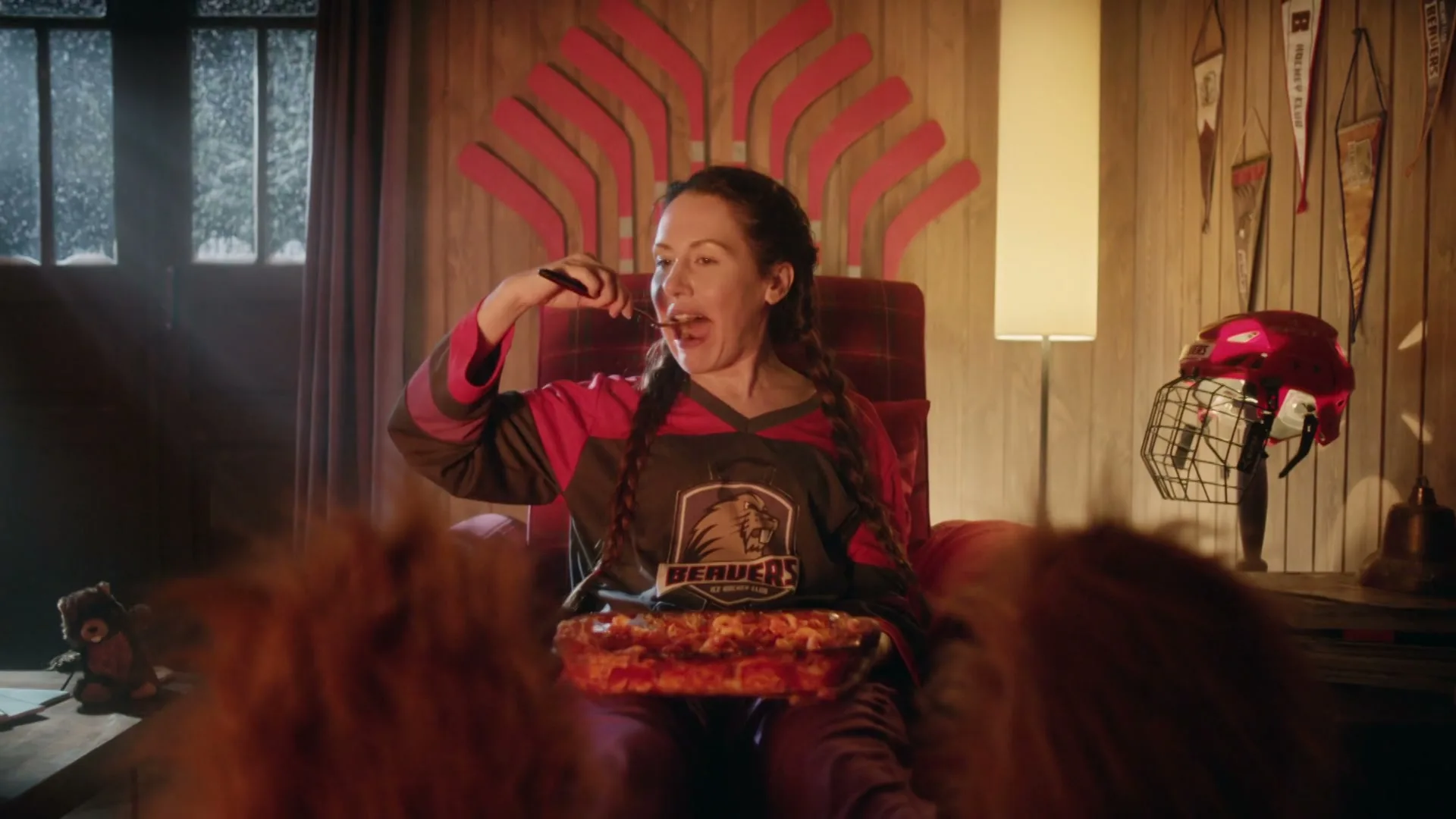 A young woman with braided hair, wearing a hockey jersey, sits in a comfy chair, eating a casserole from a glass dish in a rustic cabin.