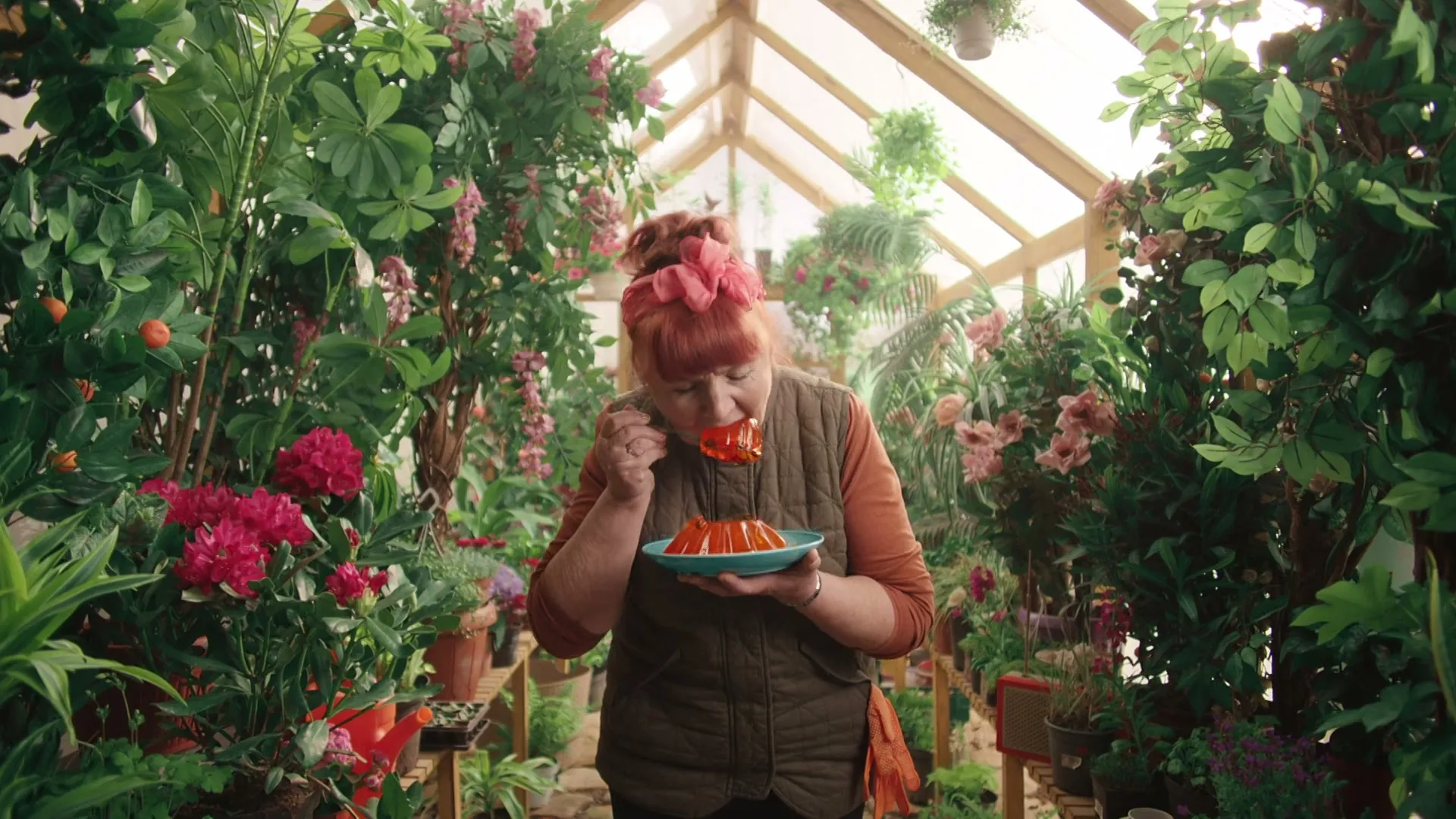 A woman with red hair and a pink bow eats orange jelly from a blue plate in a greenhouse filled with lush green plants and pink flowers.