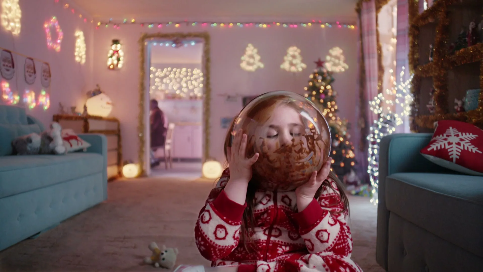 A young girl in Christmas pajamas sits on the floor, holding a clear bowl of chocolate mousse over her face in a festive living room.