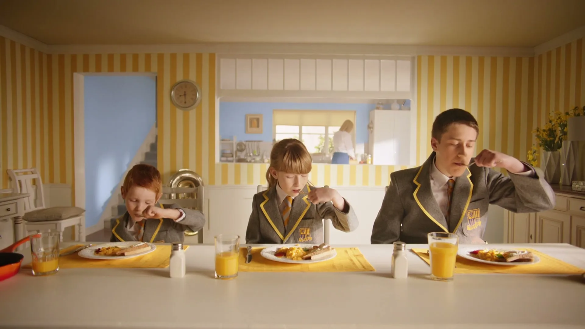 Three children in school uniforms eat breakfast at a long table, while a woman is seen in the background through a serving hatch in a brightly lit kitchen.