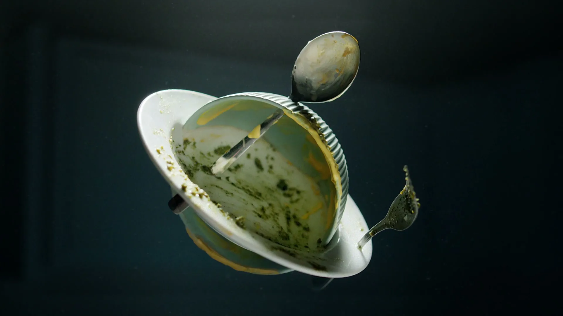 A dirty bowl, plate, spoon, and fork are spinning in mid-air against a dark background, with green food residue on them.
