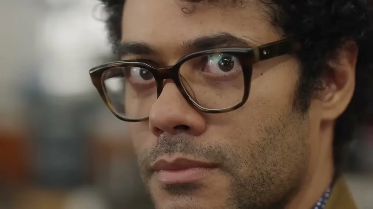 A man with curly hair and glasses looks directly at the camera, with reflections visible in his lenses, in an interior setting.