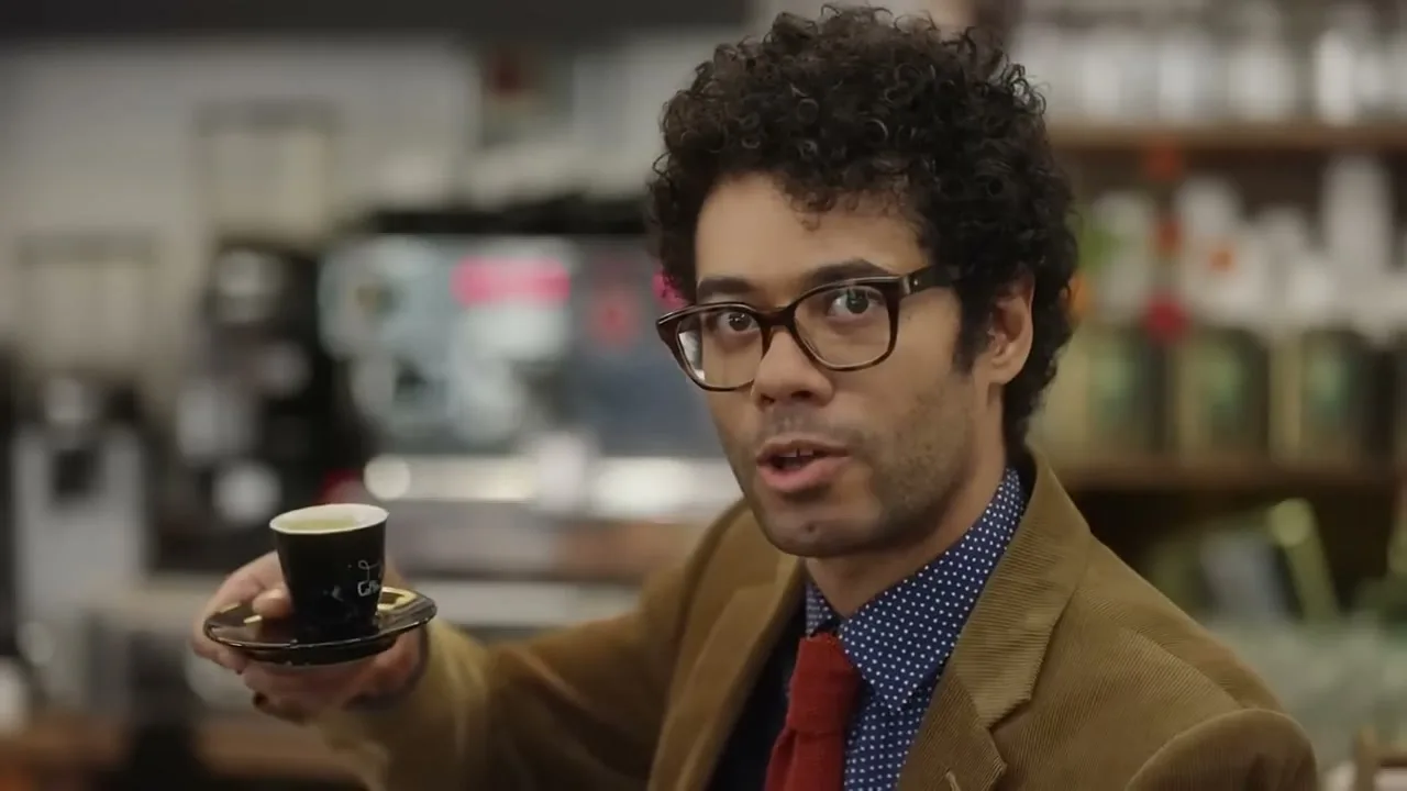 A man with curly hair and glasses holds a small cup of coffee and speaks to the camera in a cafe.
