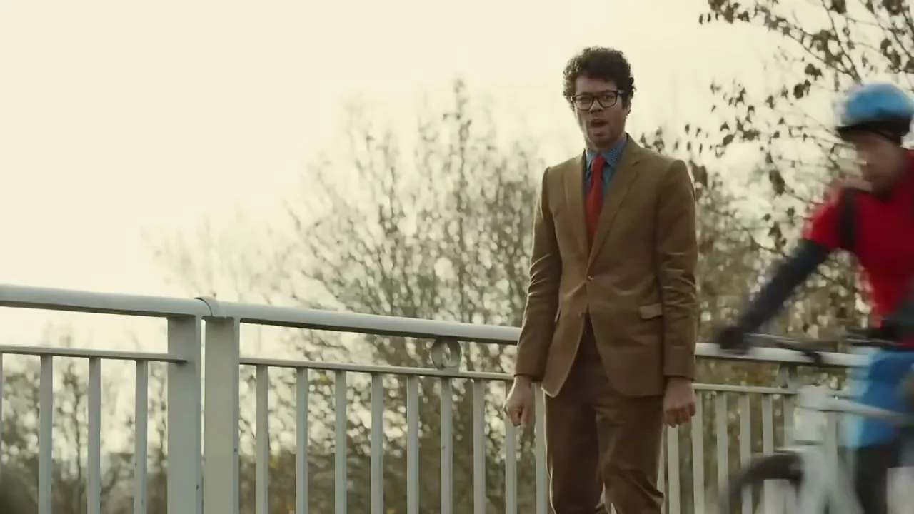Richard Ayoade, a man in a brown suit and red tie, stands on a bridge as cyclists blur past him on a cloudy day.