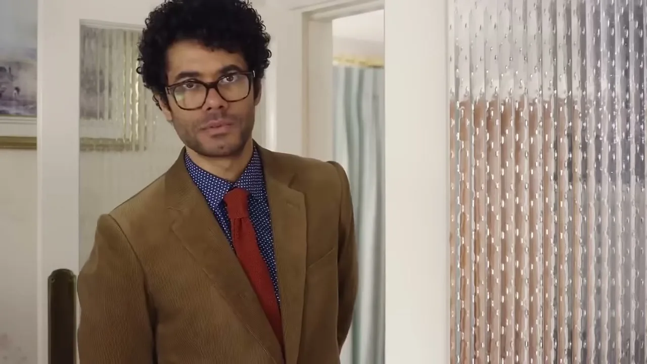 A man with curly hair and glasses, wearing a suit, looks at the camera and talks in an interior setting.
