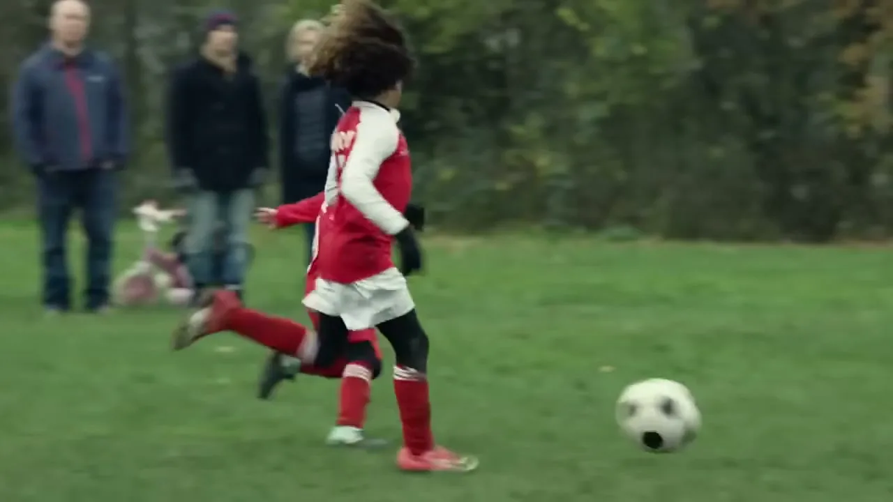 Two young boys in red soccer uniforms run on a grassy field, kicking a soccer ball during a game.