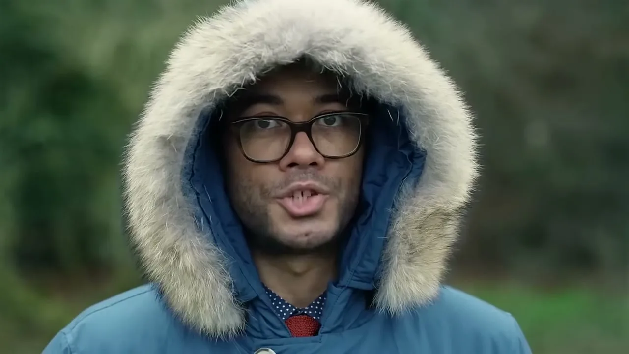 A man in his 30s wearing a blue parka with a fur-lined hood and glasses speaks directly to the camera in an outdoor setting.