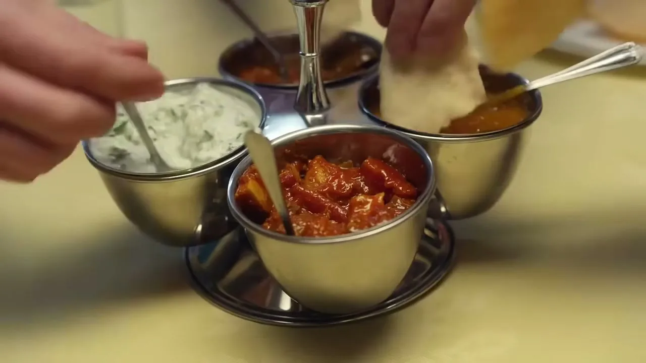 A close-up shot shows two people's hands dipping bread and a spoon into four small metal bowls of Indian chutneys and raita on a table.