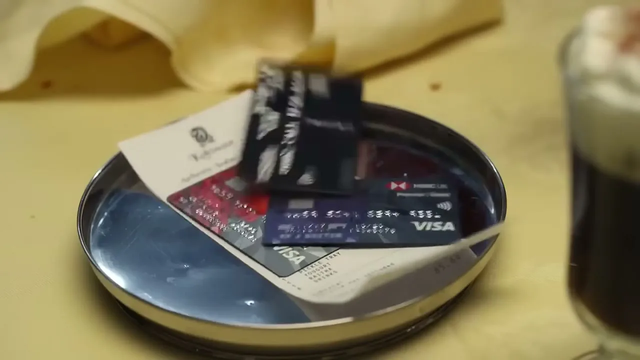 A close-up shot shows a restaurant bill on a metal tray, then multiple credit cards are dropped onto the bill, on a yellow tablecloth.