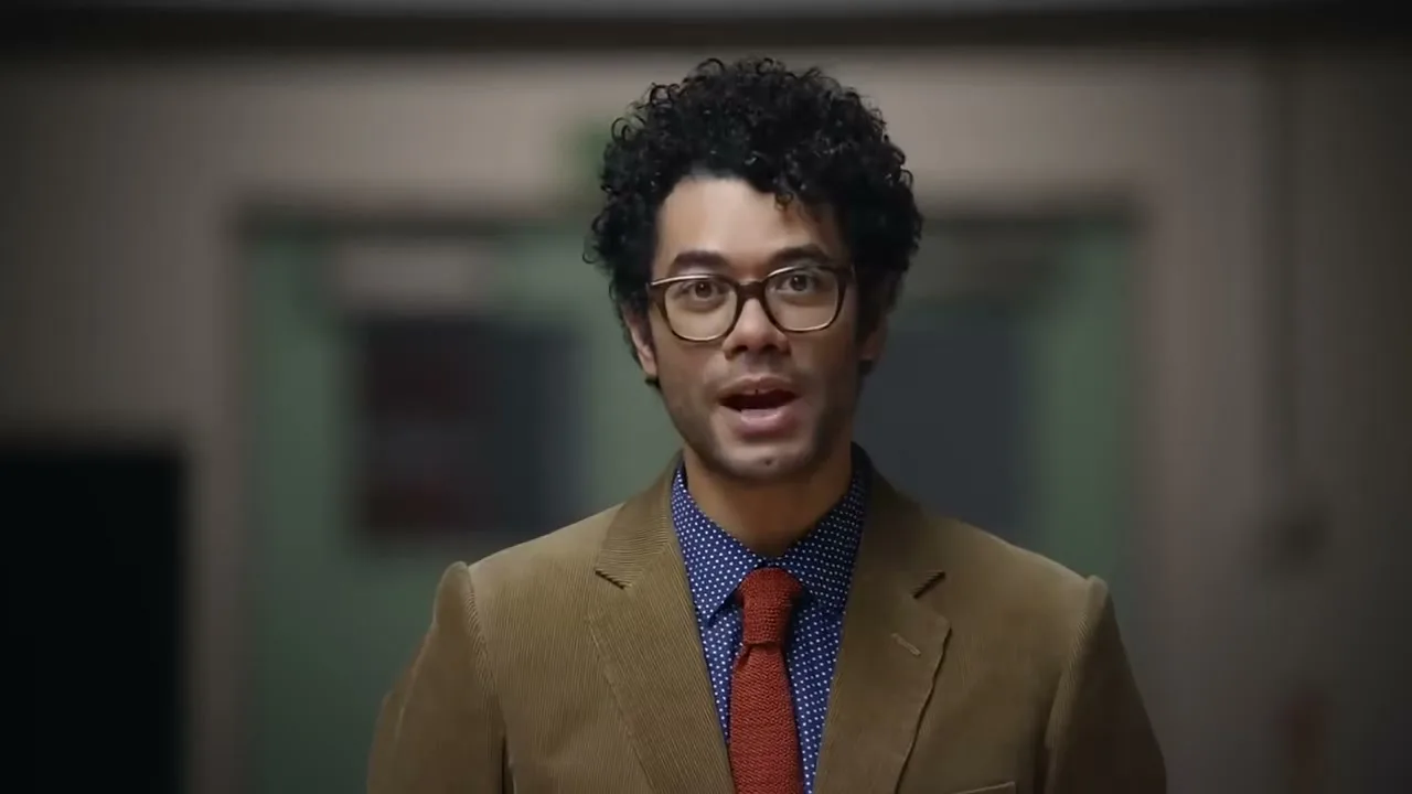 A man with curly hair and glasses, wearing a brown corduroy jacket, blue polka dot shirt, and red tie, speaks directly to the camera in an interior setting.