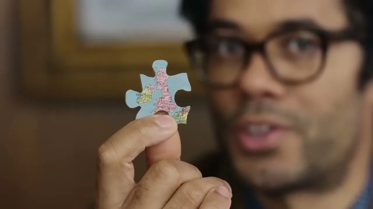 A man with glasses holds up a small puzzle piece of the UK map, speaking to the camera in an interior setting.