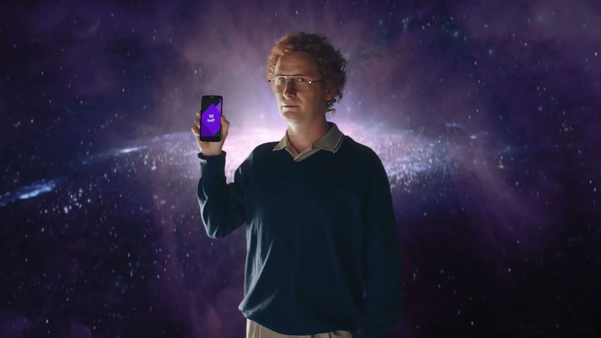 A man with curly red hair and glasses holds up a smartphone with a purple app icon, standing in front of a swirling galaxy background.