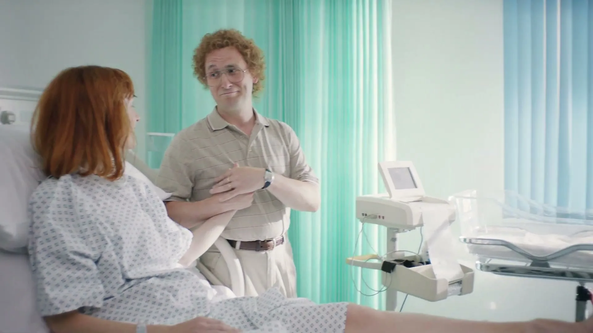 A woman in a hospital gown lies in a bed, holding hands with a man in a polo shirt, as he looks at a baby in a bassinet in a hospital room.