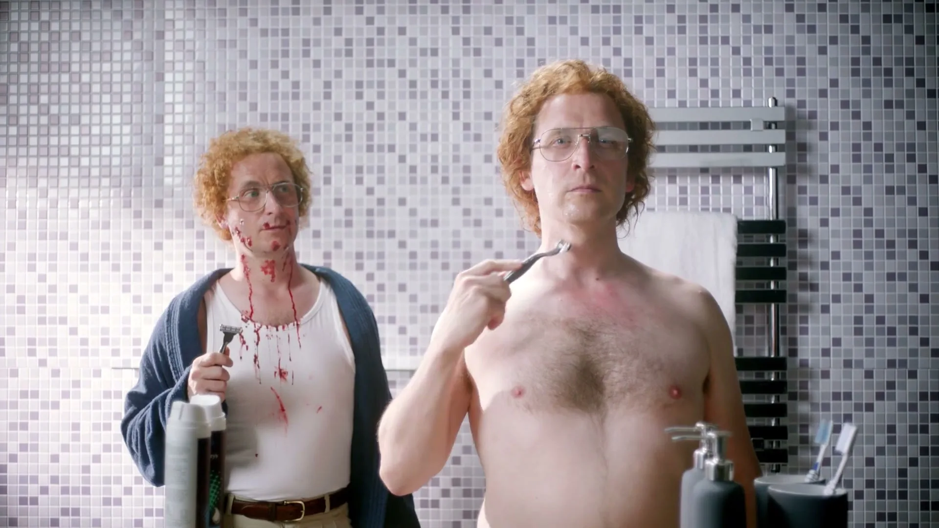 Two men with red curly hair and glasses stand in a bathroom, one shaving and the other covered in shaving cuts and blood.
