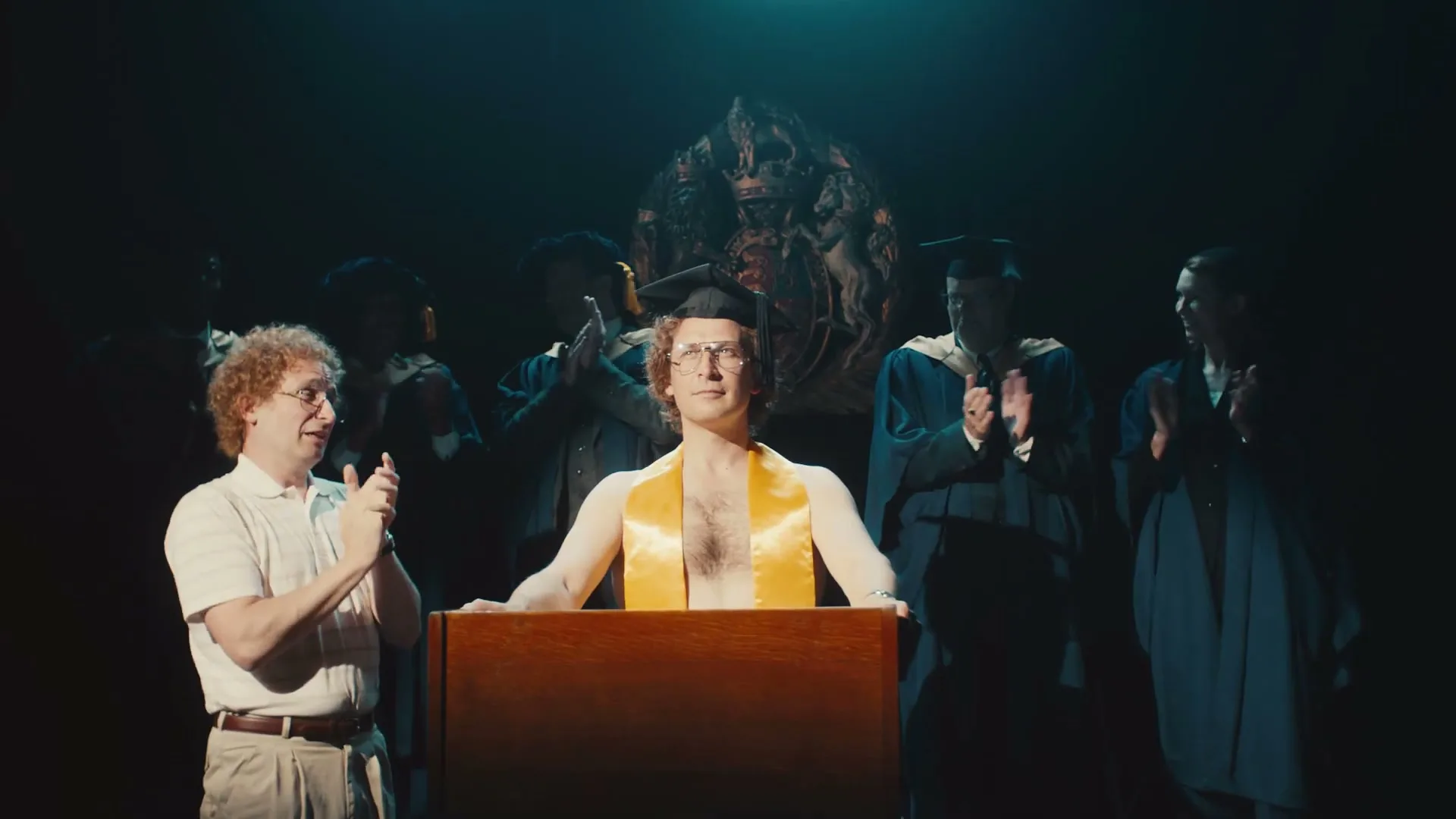 A man in a graduation cap and yellow sash, shirtless, stands at a podium while another man claps beside him, surrounded by other graduates in a dark hall.