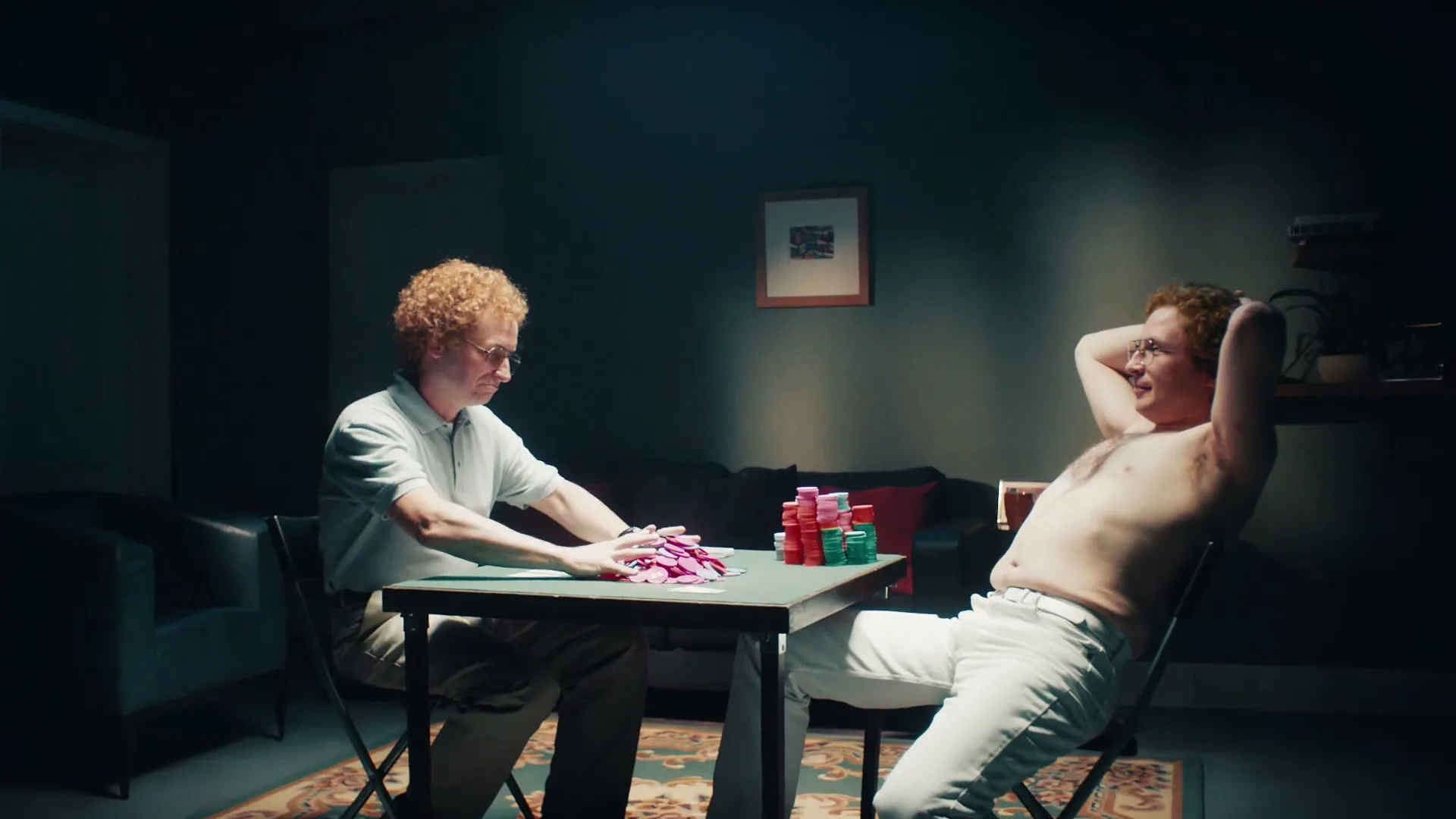 Two men with curly red hair play poker at a small table in a dimly lit room, one shirtless and relaxed, the other focused on his cards.