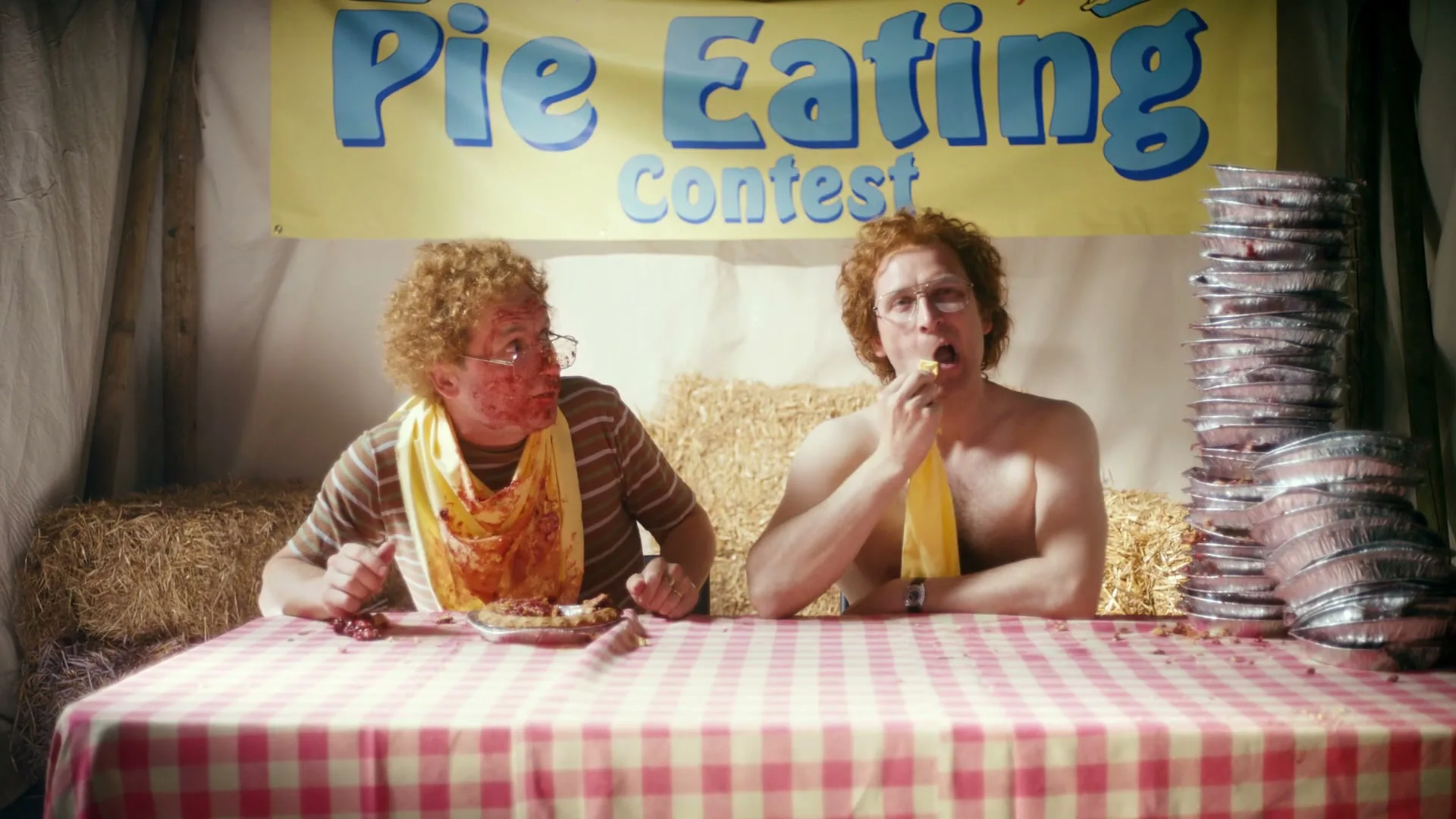 Two men with red curly hair are at a pie-eating contest, one with a messy face and the other eating a pie, at a fairground.