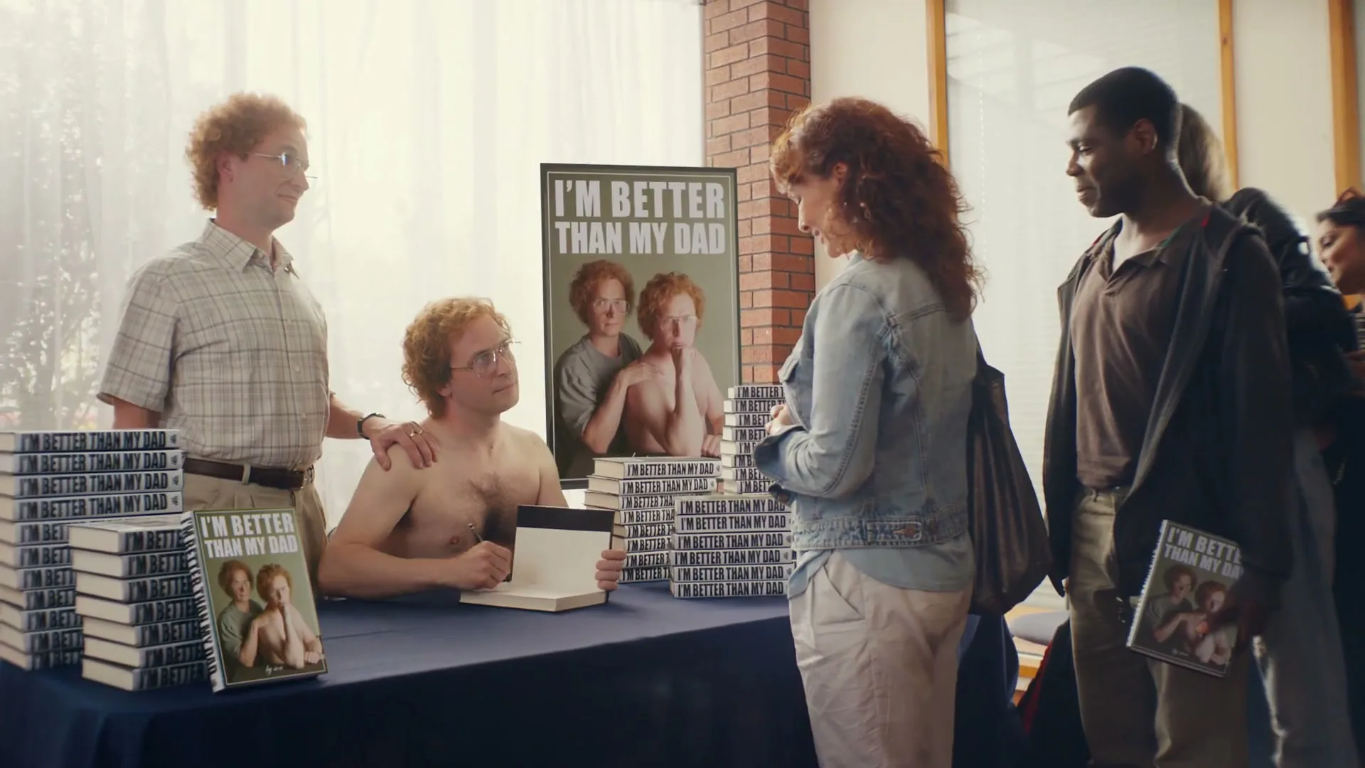 A shirtless man with red curly hair signs a book for a woman at a book signing event, while an older man with similar hair stands behind him.