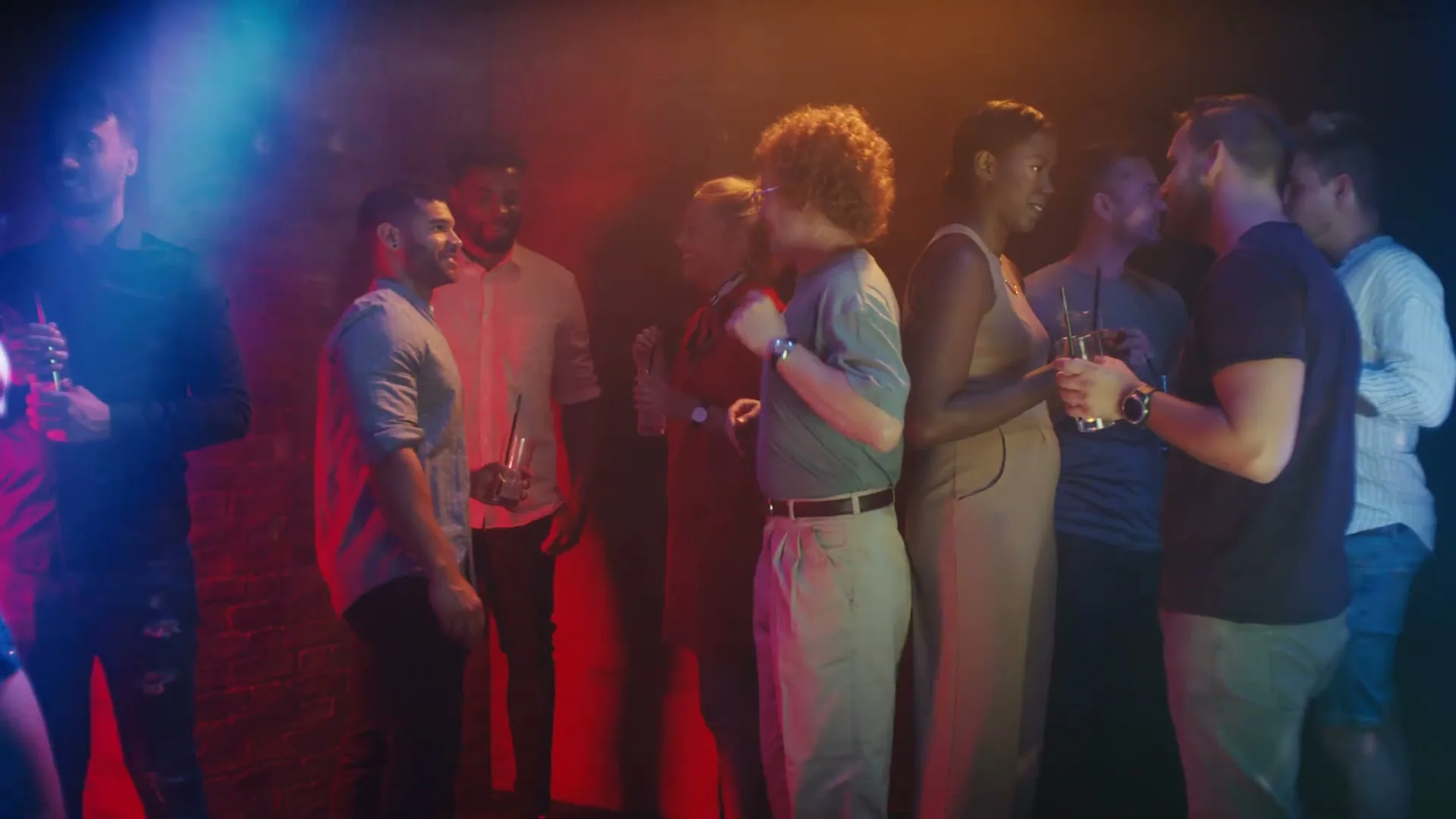 A diverse group of young adults socializes and dances at a lively nightclub, illuminated by vibrant red and blue lights.